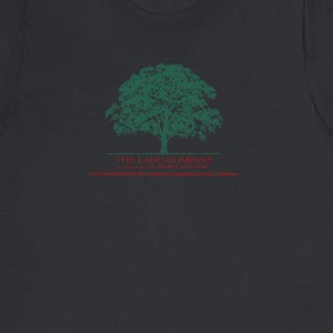 Könnte beinhalten: Dunkelgraues T-Shirt mit einem grünen Baum-Design und dem Text "THE LADO COMPANY" sowie zusätzlichem roten Text. Das T-Shirt besteht aus weichem Material und hat kurze Ärmel.