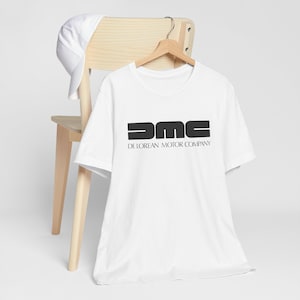 Puede incluir: Camiseta blanca con un logotipo negro que dice "DMC De Lorean Motor Company".