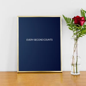 以下が含まれることがあります： 金色のフレームと濃い青色の背景の額入りプリント。「EVERY SECOND COUNTS」の文字がプリントの中央に配置されています。透明なガラスの花瓶には、赤いバラと緑の葉が入っています。