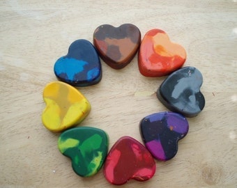 May include: A selection of heart-shaped crayons in diverse colours, such as blue, brown, orange, grey, purple, red, green, and yellow. The crayons are arranged in a circle on a light wooden surface.