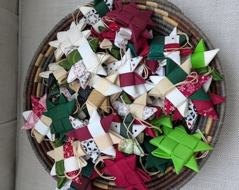 Puede incluir: Una cesta llena de adornos hechos a mano con forma de estrella. Los adornos son de tela en varios colores, incluyendo rojo, verde, blanco y beige, con detalles estampados. Cada estrella tiene una cuerda para colgar. La cesta es marrón y beige.