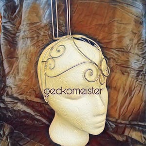 May include: A gold wire headpiece with a tall, cylindrical cage on top. The headpiece has a delicate, swirling design and is displayed on a white mannequin head.
