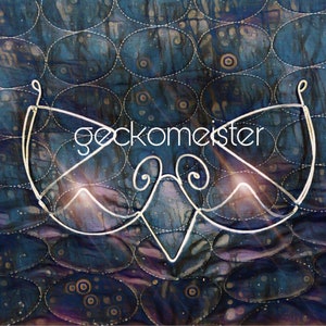 Puede incluir: Una máscara de alambre plateado con la palabra "gecko meister" escrita en ella. La máscara está sobre un fondo de tela con estampado azul y morado.