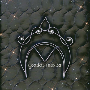 May include: A silver wire crown with a teardrop-shaped top and intricate swirls on a dark gray quilted background with sparkling glitter.