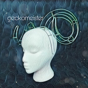 May include: A white mannequin head with a wire headpiece in the shape of ram horns. The horns are a light blue and green color. The background is a dark blue with a pattern of circles.
