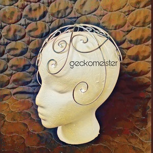 May include: A silver wire headpiece with a swirling design. The headpiece is on a white mannequin head. The text "geckomeister" is visible on the headpiece.