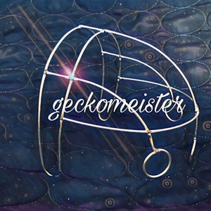 May include: A silver metal cage with a circular opening at the bottom. The cage is on a blue and white patterned fabric. The text "gecko meister" is written in white on the cage.