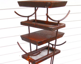 May include: A rusty metal bird feeder with four tiers and three legs. The feeder has a mesh bottom on the lowest tier.