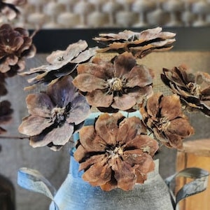 May include: A rustic metal milk jug with a distressed finish, decorated with a burlap ribbon and a bouquet of dried pine cone flowers.