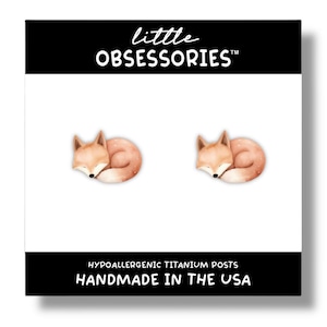 May include: A pair of hypoallergenic titanium post earrings featuring a sleeping fox design. The fox is curled up in a ball with its tail wrapped around its body. The earrings are handmade in the USA. little OBSESSORIESTM