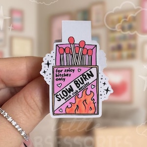 May include: A pink and white sticker in the shape of a matchbox, with the text "SLOW BURN book club matches" and "for spicy bitches only." The sticker features a flame graphic and red match heads.