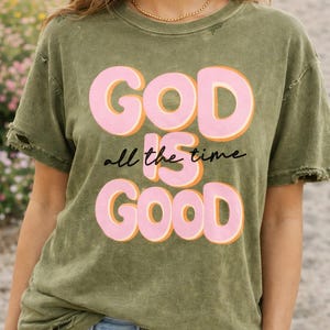 Puede incluir: Camiseta verde oliva con las palabras "GOD is all the time GOOD" en letras rosas con contornos naranjas. La camiseta tiene un aspecto desgastado con bordes deshilachados y se combina con vaqueros azules.