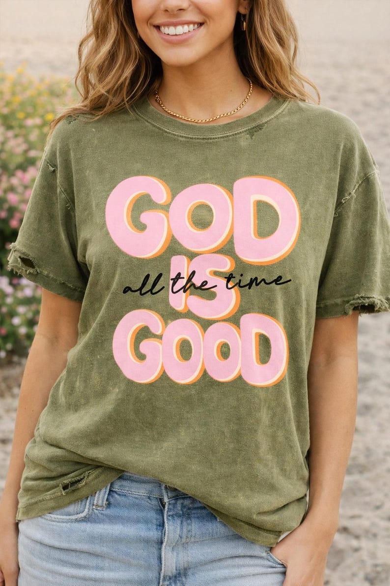 Puede incluir: Camiseta verde oliva con las palabras "GOD is all the time GOOD" en letras rosas con contornos naranjas. La camiseta tiene un aspecto desgastado con bordes deshilachados y se combina con vaqueros azules.