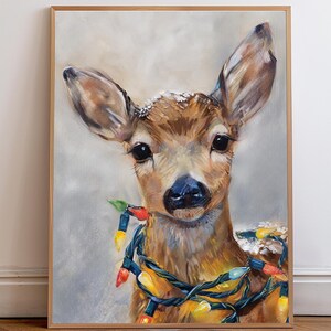 May include: A framed painting of a deer with large ears and dark eyes, adorned with a string of colorful Christmas lights. The deer has a brown coat and a blue nose, with a light gray background.