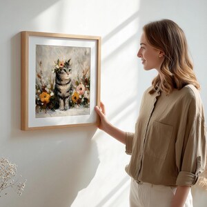 Puede incluir: Una impresi&oacute;n art&iacute;stica enmarcada que presenta un gato atigrado con una corona floral, rodeado de flores. La obra de arte tiene un estilo suave y pict&oacute;rico con un fondo neutro y un marco de madera clara. El gato es el foco principal de la imagen.