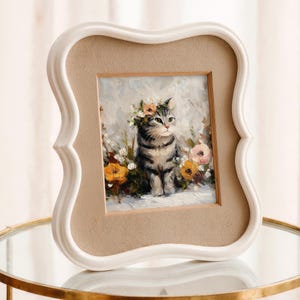 Puede incluir: Un retrato enmarcado de un gato. La pintura representa un gato atigrado gris con una corona de flores, rodeado de flores coloridas. El marco es blanco con un borde decorativo y un paspart&uacute; beige.