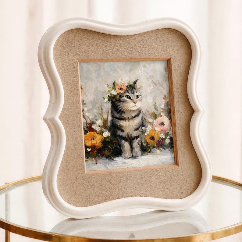 Puede incluir: Un retrato enmarcado de un gato. La pintura representa un gato atigrado gris con una corona de flores, rodeado de flores coloridas. El marco es blanco con un borde decorativo y un paspart&uacute; beige.