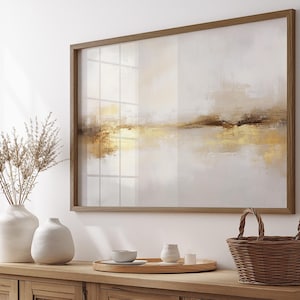 May include: Abstract landscape art print in a wooden frame. The artwork features a blend of neutral tones, including white, beige, and gold, creating a serene and modern aesthetic. The framed art is displayed above a wooden cabinet with decorative items.