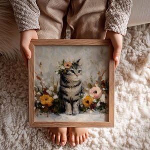 Puede incluir: Pintura enmarcada cuadrada de un gato atigrado gris con una corona floral, rodeado de flores coloridas. La obra de arte tiene un estilo suave y pict&oacute;rico con un fondo claro. El marco es de madera clara.