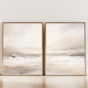 Puede incluir: Dos pinturas de paisajes abstractos enmarcadas. La obra presenta una paleta de colores neutros de beige, gris y blanco, con un efecto suave y borroso. Las pinturas están en marcos de madera marrón claro, creando un conjunto cohesivo para la decoración del hogar.