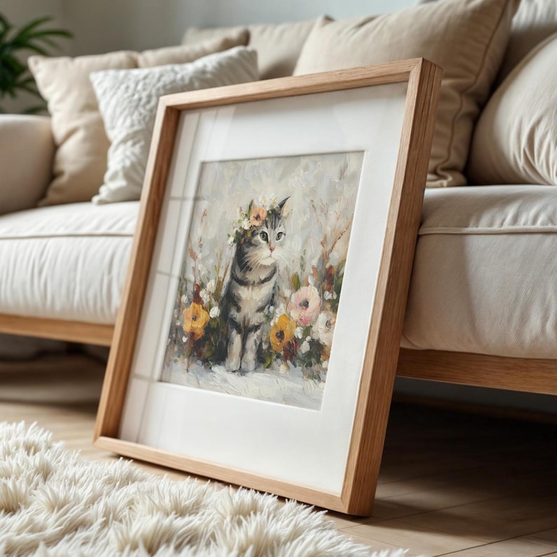 Puede incluir: Una impresi&oacute;n art&iacute;stica enmarcada que presenta un gato atigrado con una corona de flores, rodeado de flores coloridas. La obra de arte est&aacute; enmarcada en madera con un passe-partout blanco, apoyada contra un sof&aacute; crema. El gato es gris y blanco.