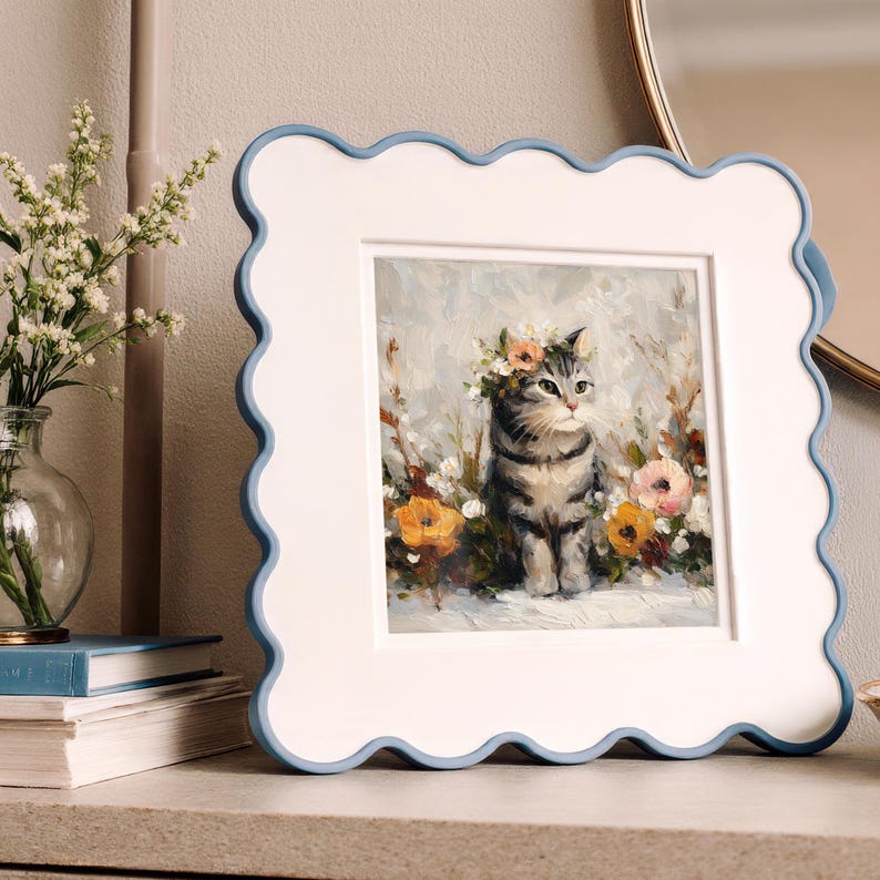 Puede incluir: Cuadro enmarcado de un gato atigrado gris con una corona de flores, rodeado de flores. El marco es blanco con un borde festoneado azul claro. La obra de arte se exhibe sobre una superficie neutra.