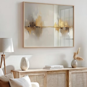 Puede incluir: Una pintura abstracta enmarcada con una escena de paisaje horizontal en tonos beige, marrón y dorado. La pintura está colgada en una pared blanca sobre un armario de madera.