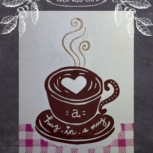 May include: A brown coffee mug with a heart design and steam rising from it. The mug is on a pink and white checkered background with the text "a= hug. in. a mug" underneath. The text "COFFEE" is at the bottom left and "MILK FLAVOR LATTE" is at the bottom right.