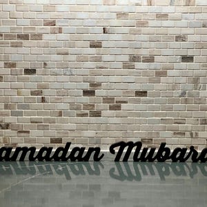 May include: Black wooden sign with the words "Ramadan Mubarak" in a cursive font. The sign is placed on a reflective surface in front of a brick wall with a mix of white, gray, and brown bricks.