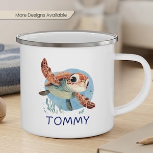 May include: White enamel mug with a cartoon sea turtle design. The turtle is brown, orange, and green, with the name "TOMMY" in blue below. The mug has a silver rim and a white handle. Text at the top reads "More Designs Available."