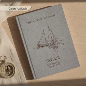 Puede incluir: Un diario gris con el título "THE ADVENTURES OF" y una ilustración de un velero. El nombre "TAYLOR" está impreso debajo de la ilustración, junto con la ubicación "Bali, Indonesia" y la fecha "20 de junio de 2026". Una brújula es visible en la esquina inferior izquierda.