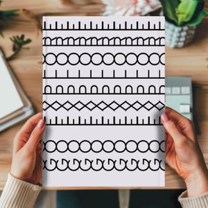 Puede incluir: Un cuaderno blanco con un patrón geométrico negro de líneas, círculos y formas. El cuaderno está sostenido por una persona. El diseño se repite en la portada. El cuaderno está sobre una superficie de madera.