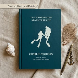 Könnte beinhalten: Ein dunkelblaugrünes Hardcover-Buch mit einer weißen Grafik von zwei Tauchern. Der Text lautet: THE UNDERWATER ADVENTURES OF, CHARLIE & JORDAN, MANTA POINT, 04 10.000' N, 73 30.000. Das Buch ist mit einer Muschel, einer Fächerkoralle und einem Stift arrangiert.