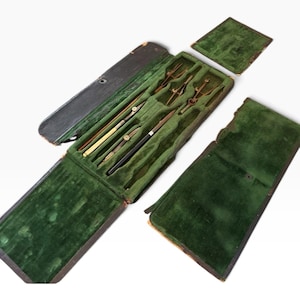 May include: A vintage drafting instrument set housed in a dark brown case with a green velvet interior. The set includes several brass and metal compasses and drawing tools arranged in molded slots, with a separate matching green velvet lid.