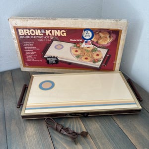 Puede incluir: Bandeja eléctrica caliente Broil King Deluxe vintage. La bandeja es blanquecina con rayas marrones, azules y beige y un círculo azul y beige. Tiene asas marrones y un cable de alimentación marrón. La caja es visible en el fondo.