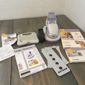 May include: A collection of Xyron scrapbooking tools and accessories, including a label maker, design runner, and various cartridges. The items are white and purple, with printed instructions and packaging. The products are arranged on a wooden surface.