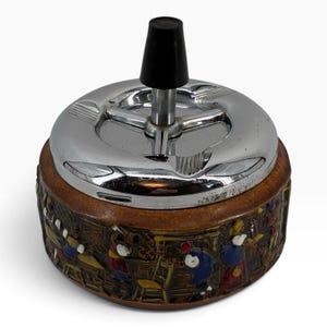 May include: A round spinning ashtray with a polished chrome metal top and a black plastic handle. The base is made of brown ceramic featuring a relief-molded scene of figures in colorful clothing engaged in various activities around a room.