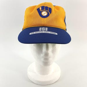 Könnte beinhalten: Vintage Baseballkappe mit goldfarbener Krone und blauem Schirm. Die Kappe zeigt ein Baseballhandschuh-Logo mit einem Baseball in der Mitte. Auf dem Schirm steht der Text "THE OFFICIAL SOFT DRINK".