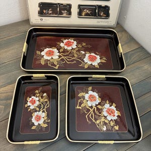 Puede incluir: Un juego de tres bandejas negras para servir con un diseño floral. Las bandejas tienen un fondo rojo oscuro con flores blancas y rojas y detalles dorados. Las bandejas tienen asas tejidas y ribete dorado.