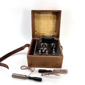 Puede incluir: Un antiguo dispositivo médico Sands Battery No. 10 en una caja de madera con una correa de cuero. La caja está abierta, revelando componentes internos y un documento impreso. Dos electrodos cilíndricos con mangos negros y cables están delante.