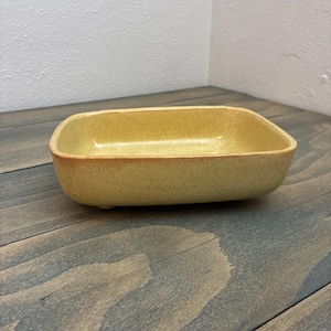 May include: A rectangular ceramic dish with rounded corners. The dish is a speckled yellow color with a slightly darker rim. The interior is smooth, while the exterior has a textured appearance. The dish is sitting on a wooden surface.