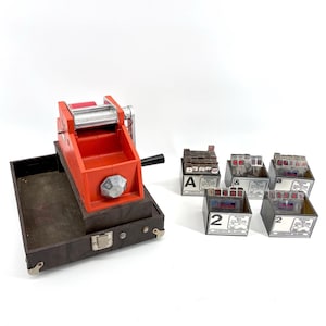 May include: A vintage red Lettertype machine with a black handle and a brown case. Several small boxes with letters and numbers are also present. The machine has a silver roller and a dial with numbers.