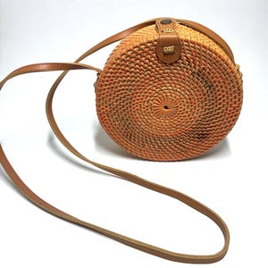 May include: A round, woven rattan crossbody bag in shades of orange and brown. The bag features a brown leather strap and a leather closure. The bag is approximately 8 inches in diameter.