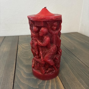 May include: A red candle with a carousel design. The candle features detailed carvings of figures, including children and adults, in a circular arrangement. The candle has a conical top and is made of a solid, red wax.