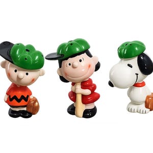 May include: Three ceramic figurines of Peanuts characters in baseball attire. Charlie Brown wears an orange shirt and green cap. Lucy holds a bat, and Snoopy has a baseball glove. All wear green baseball caps.