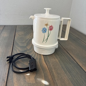May include: Off-white electric coffee pot with a floral design in blue, red, and gold. The pot has a lid, handle, and spout. A black power cord is next to the pot. The pot is on a wooden surface.