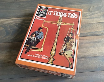 May include: A vintage board game box for "IT TAKES TWO" by Hasbro. The box has a red background with a gold scale graphic and illustrations of couples. The text "THE EXCITING NEW COUPLES GAME" is also visible.
