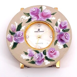 Puede incluir: Un reloj de pulsera redondo con una esfera beige pintada a mano con rosas moradas y hojas verdes. La esfera presenta un bisel dorado, cuatro pequeñas piedras transparentes como marcadores de hora, manecillas doradas y el texto Marie Lourdes y Quartz.