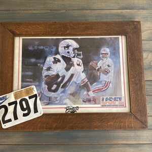 Op de afbeelding: Ingelijste prent van twee American football-spelers in witte en bruine uniformen. De prent zit in een bruine houten lijst. Een wit en zwart Nike-label met het nummer 2797 staat in de linkerbenedenhoek.