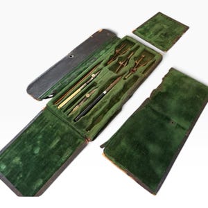 Puede incluir: Un juego de instrumentos de dibujo técnico en un estuche rectangular de color marrón oscuro con interior de terciopelo verde. El set incluye varios compases y divisores de latón y metal, además de una herramienta con mango negro en compartimentos.